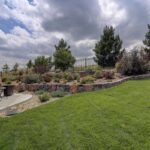 How to Create Fire-Resistant Landscaping Backyard with green lawn, stone retaining wall, drought-tolerant landscaping, patio seating, and fire-resistant plants under partly cloudy sky.