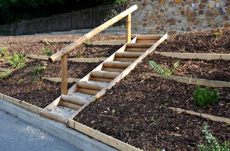 Wooden log stairs with gravel steps and a handrail ascend a terraced dark mulch slope planted with small green shrubs.