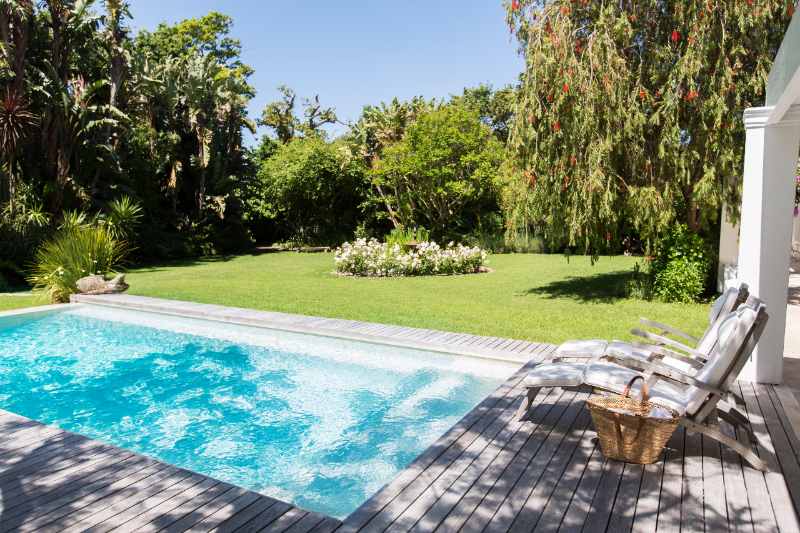 Lawn chairs in the shade of a tree on a wooden deck overlooking a backyard swimming pool and lush green lawn