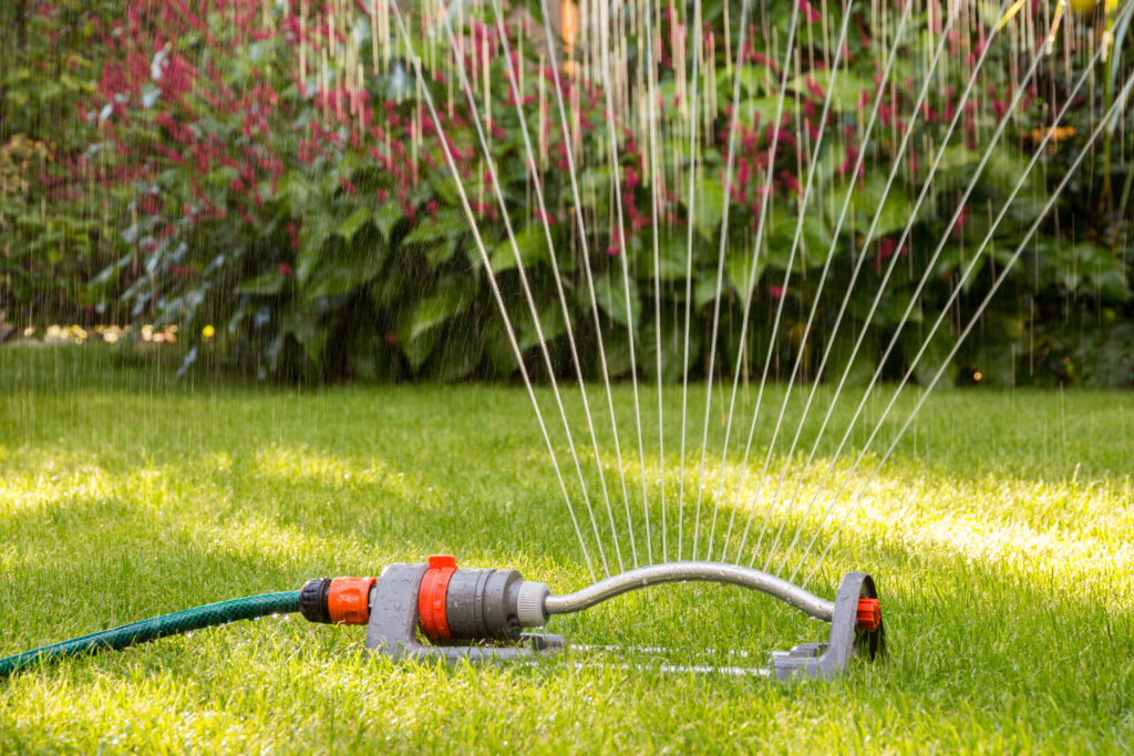 An oscillating lawn sprinkler spraying arching streams of water over a lush green lawn with colorful garden plants in the background.