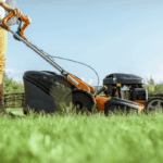 How to Know When to Stop Mowing for the Season A man in yellow pants pushes an orange gasoline-powered lawn mower across a green lawn on a sunny day, with a wooden fence and trees blurred in the background.