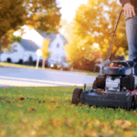 7 Fall Lawn Care Mistakes That Cost Homeowners Come Spring A person mows a suburban lawn with fallen autumn leaves during.