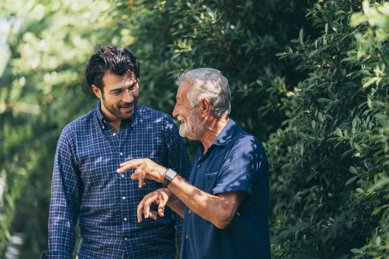 An older gray-haired man in a blue shirt gestures as he talks with a smiling younger man in a plaid shirt, with lush green hedges behind them in a sunlit backyard.
