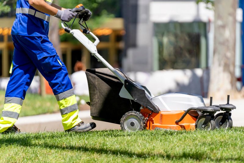 A professional lawn care worker in a blue uniform with reflective yellow safety stripes pushes an orange and gray bagging lawn mower across a sunlit green lawn in a park.