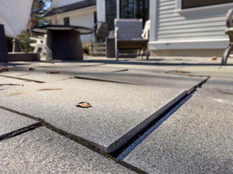 A low-angle view of a gray stone patio where one paver has lifted above its neighbors, leaving a raised edge and gap, with patio chairs and a house in the background.
