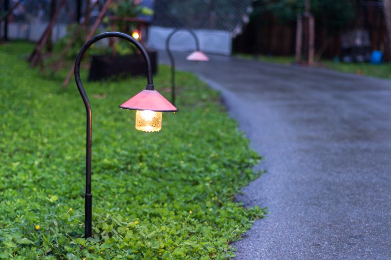 Two curved-post walkway lights with pink conical shades glow warmly along a wet paved path at dusk, with a green groundcover lawn beside the path.
